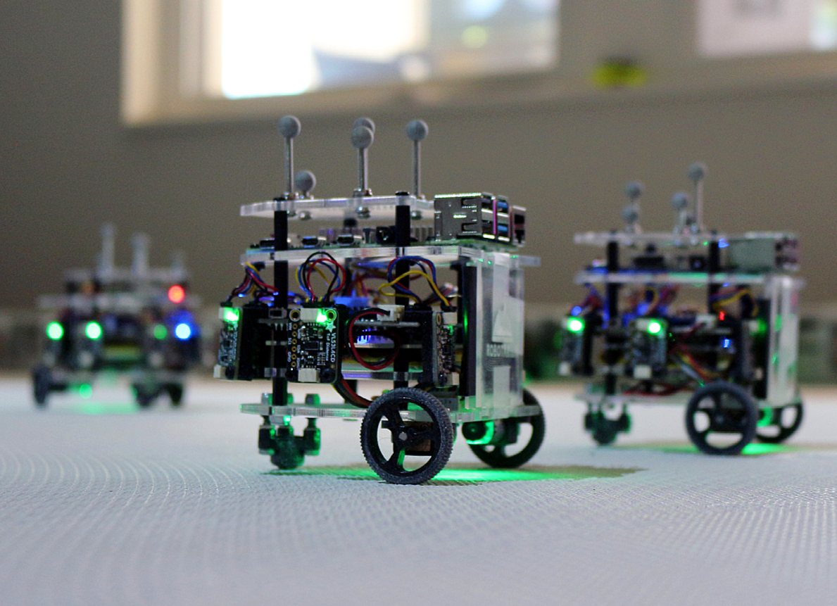 small box like robots on wheels on a table top. Some are in foreground and others in the background.